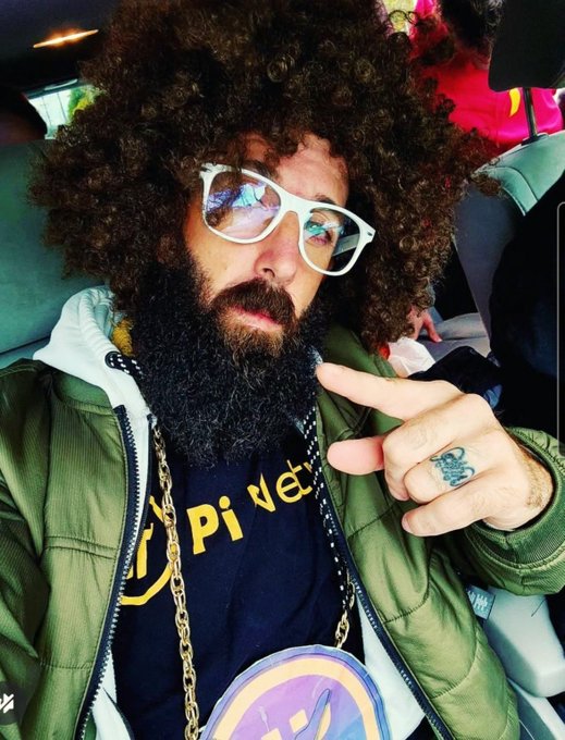 A man with curly hair, beard, white-framed sunglasses, gold chains, and a green jacket makes a peace sign while wearing a black 'private' t-shirt.