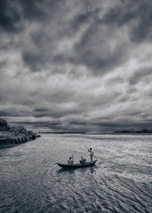 A black-and-white photograph showing three people in a small boat rowing on a wide river under a dramatic, cloudy sky, with distant shoreline visible on the horizon.