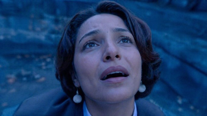 A close-up shot of a woman with short dark hair wearing pearl drop earrings, looking upward with an emotional, tearful expression against a dim blue-lit background.