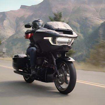 A motorcyclist in a helmet rides a black touring motorcycle down a road with mountains in the background.