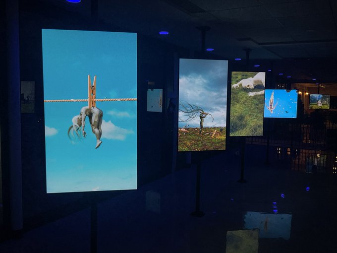 An array of illuminated digital screens in a dark gallery space displays surreal artwork, with the most prominent screen featuring a figure suspended upside down by their feet from a clothespin attached to a clothesline against a blue sky—a literal interpretation of "hanging in there." Additional screens show imaginative scenes including a streaking comet and floating figures, all presented as part of a digital art exhibition at 0.3 ETH.