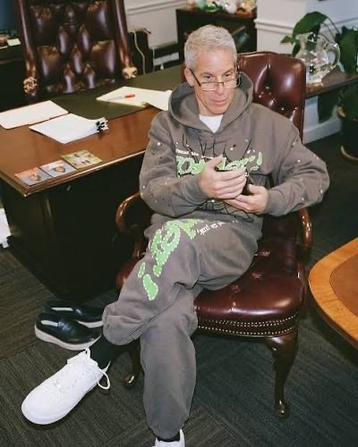 A man with short bleached hair and glasses, wearing a gray matching sweatshirt and sweatpants with green graphic designs, sits in a dark red leather chair holding a phone, with white sneakers on the floor.