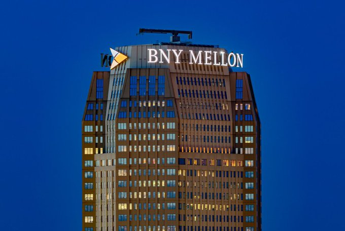 The image shows the illuminated "BNY MELLON" logo atop a skyscraper against a deep blue evening sky. The visual ties directly to the news of the historic $2.2 trillion bank increasing its Bitcoin exposure through additional purchases of MicroStrategy ($MSTR) shares.