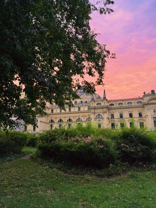 A large ornate stone building stands behind lush green bushes and trees, with a pink and purple sunset sky in the background.