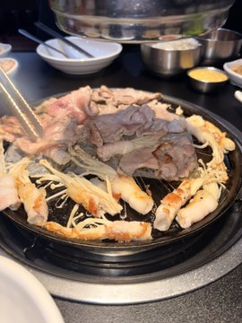 A Korean BBQ grill loaded with raw beef slices, enoki mushrooms, and shrimp, surrounded by small side dishes (banchan).
