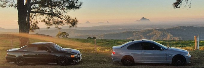 Two sedans, a black one and a silver one, parked facing each other on a grassy hilltop overlooking a misty mountainous landscape during sunset.