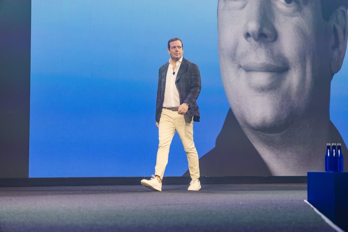 A man in a blazer and khaki pants stands on a stage during a presentation, with a large screen behind showing a blue-tinted close-up of his face and water bottles on a stand to the right.