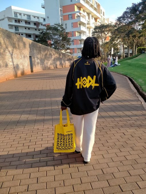 A person walks along a brick pathway wearing a black jacket with "KOOL" in yellow lettering, while carrying a bright yellow tote bag printed repeatedly with "BINANCE." The branded merchandise from the post showcases Binance swag, fitting the caption's reference to their gear collection.