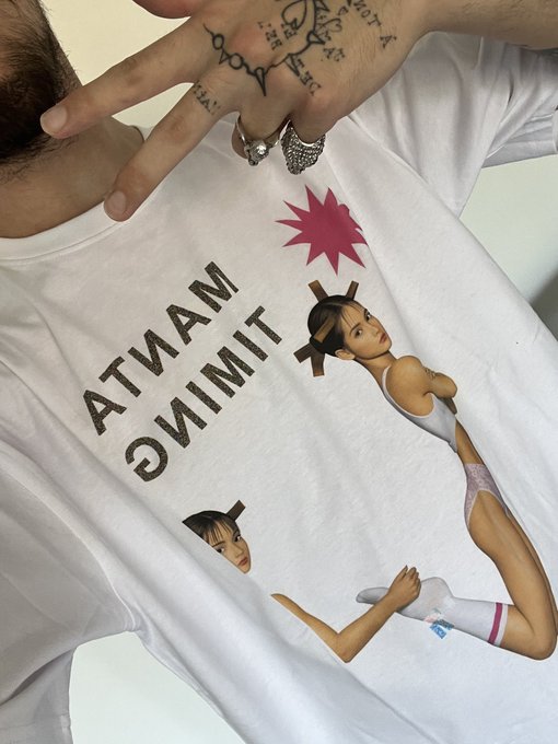 A person models a white "MANTA TIMING" graphic tee featuring anime-style illustrated characters and a pink starburst, with their tattooed hand and silver rings raised near the collar, showcasing the brand's latest drop.