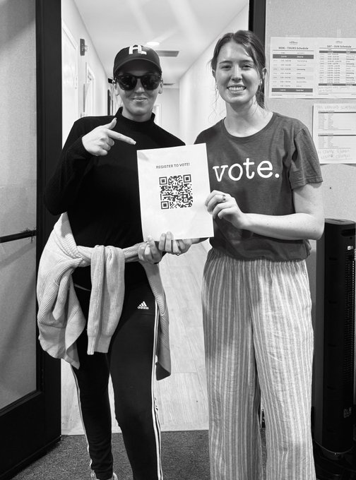 A black-and-white photo of two smiling young women standing in a hallway; one wears a cap and sunglasses, while the other wears a shirt reading 'vote.', and together they hold up a paper with a QR code and the text 'REGISTER TO VOTE!'.