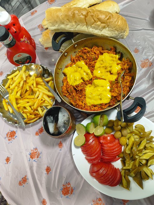 A table spread features a pan of rice topped with melted cheese, french fries, sliced tomatoes, pickled cucumbers, olives, bread, and ketchup.