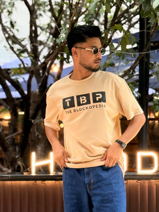 A man wearing sunglasses and a tan t-shirt featuring the 'TBP THE BLOCKOPEDIA' logo stands outdoors with trees and a neon sign in the background.