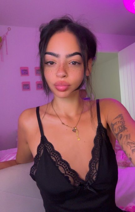 A selfie of a young woman with dark hair and freckles wearing a black lace top and gold necklaces, in a room illuminated with pink lighting.