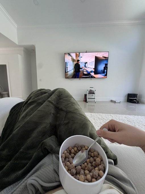 A person sits cozily on a couch under a green blanket, holding a bowl of cereal while watching TV, embodying the post's sentiment of being blissfully detached from the constant crypto chatter ("everyone going back and fourth about crypto this crypto that orangie:")—the "orangie" likely referring to their carefree, orange-shaded morning vibe of ignoring the noise in favor of a chill breakfast and screen time.