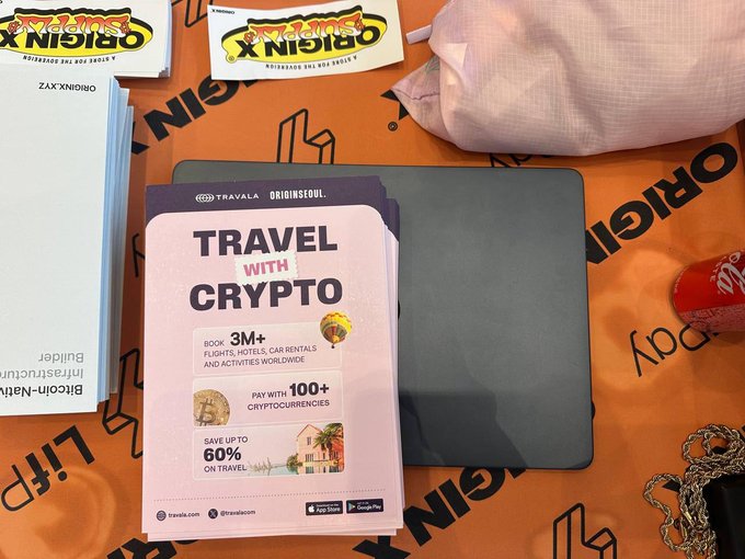A promotional travel pamphlet reading 'TRAVEL WITH CRYPTO' by Travala offering 3M+ bookings with 100+ cryptocurrencies and up to 60% savings, sitting on an orange Origins-branded table alongside a book titled 'Bitcoin-Native Infrastructure Builder'.