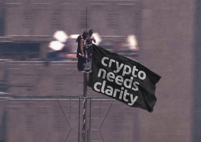 Two people stand atop a tall pole or antenna holding a large black flag that reads "crypto needs clarity," with a hazy cityscape blurred in the background. The dramatic aerial display visually amplifies the post's urgent message that the cryptocurrency industry is ready for clearer regulatory guidelines.
