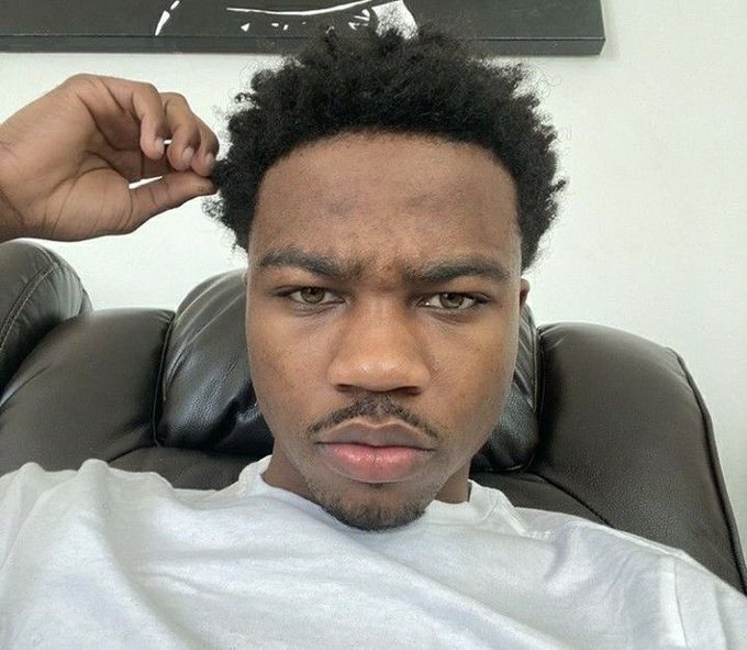 A young man with a serious expression rests on a leather couch, dressed in a white t-shirt, as he greets his followers with a calm "gm" while contemplating how to make money today.