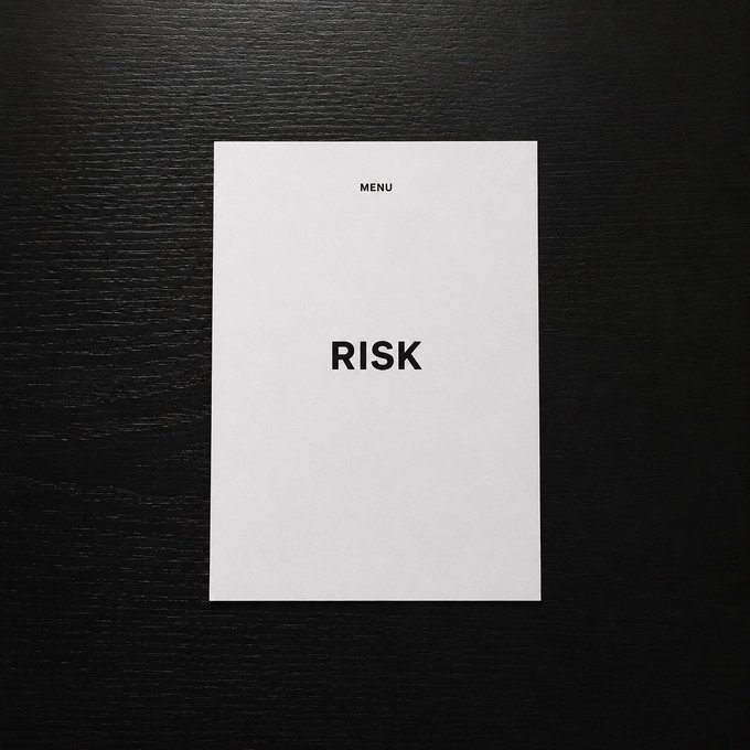 A minimalist white menu card with the text 'MENU' at the top and 'RISK' printed in the center, resting on a dark wood surface.