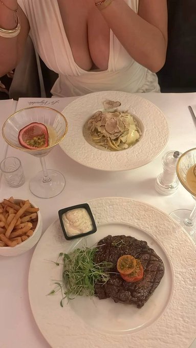A woman in a white dress sits at a restaurant table featuring a plated steak with microgreens, a pasta dish topped with shaved truffles, fries with aioli, and cocktails garnished with passion fruit. The elegant meal setup complements the appreciative "gmgm" caption, suggesting the diner is savoring a refined dining experience.