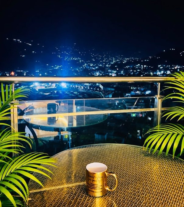 The image features a glass table with a gold coffee cup sitting on top of it, overlooking a city at night. The scene suggests that someone has left their coffee cup on the table while they were away or perhaps took a break from enjoying their drink. The photo is taken from an elevated position, providing a unique perspective and showcasing the beautiful view of the city lights below.