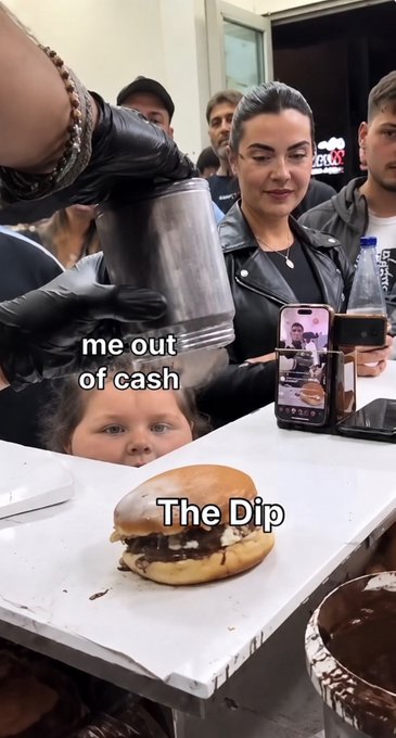 A vertical video frame showing a person in black gloves dipping a large burger on a counter while labeled 'The Dip', with a woman and child watching, captioned 'me out of cash'.