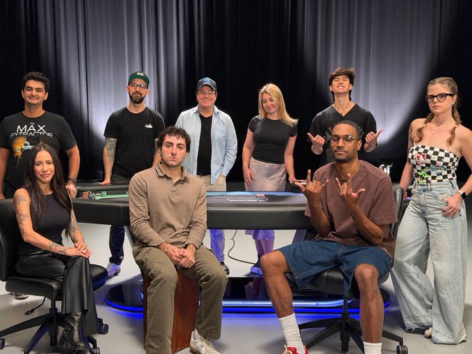 A group of nine people pose around and behind a poker table in a studio setting with dark curtains, with one man's shirt reading "MAX EVERYTHING" in visible text. The casual, posed arrangement of players at the table supports the caption's claim that the image is a real, unedited photograph rather than AI-generated.