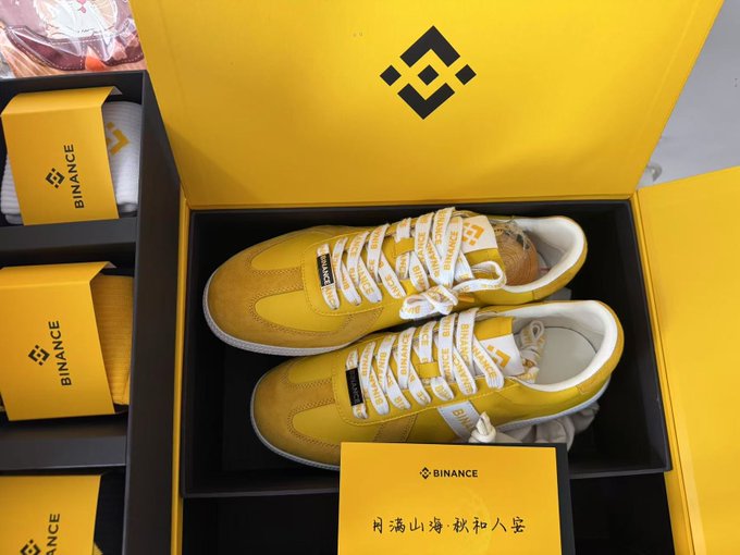 A pair of yellow and white sneakers branded with "BINANCE" logos are displayed in an open yellow gift box featuring the Binance diamond logo, with additional Binance-branded packaging visible on the side and a card with Chinese text. The image showcases the Mid-Autumn Festival gift box mentioned in the post, highlighting the matching shoes and socks as promotional merchandise.