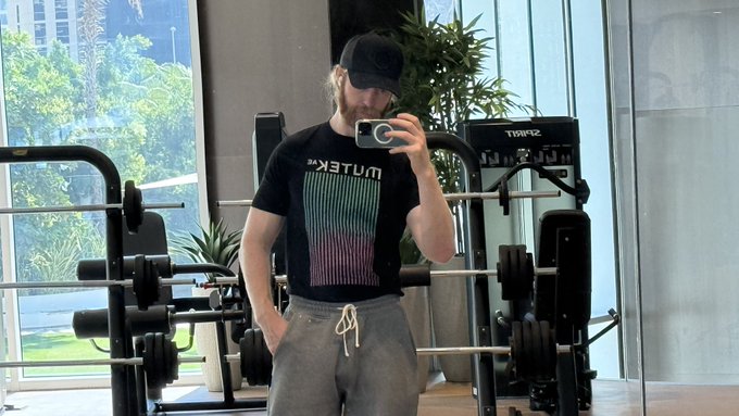 A bearded man wearing a black cap, gray sweatpants, and a black 'EXRETUM' t-shirt takes a mirror selfie with a smartphone in a gym, with weight equipment and potted plants visible behind him.