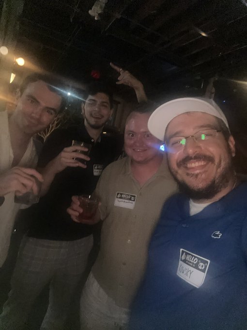 A group selfie of four men smiling in a dimly lit setting, with some wearing name tags that read 'Hello My Name Is', including one tag for 'LUSKEY' on a man in a blue shirt and white cap.