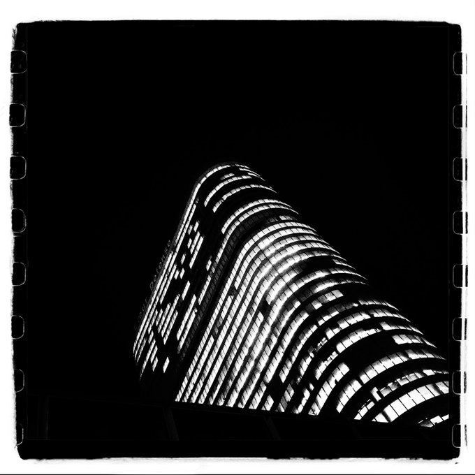 A black and white film photograph captures a modern high-rise building at night with illuminated windows, framed by a visible film border.
