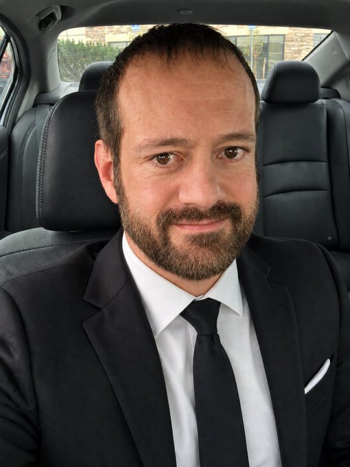 A close-up portrait of a man with short brown hair and a beard, wearing a dark suit, white shirt, and dark tie, seated in the driver's seat of a car with black leather interior.
