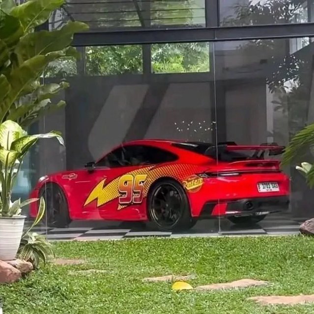 A red Porsche sports car with a 'Lightning McQueen 95' wrap is parked inside a glass-walled garage with a grassy yard in the foreground.