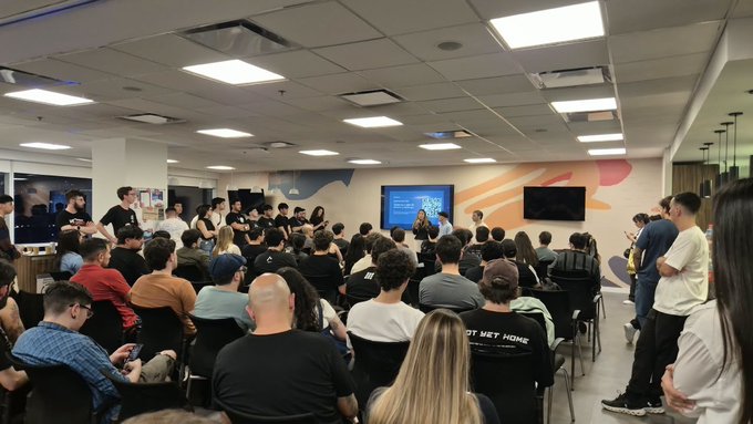 A large crowd of people sits and stands in a room watching a presentation on screens, including one displaying a world map.