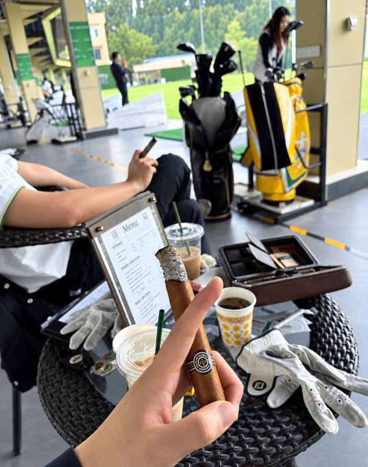 A person holds a partially smoked cigar between their fingers at a golf course lounge, with a menu, iced coffee, golf gloves, and golf bags visible on the table. The image complements the post's reflection on staying grounded emotionally while growing capital in crypto—pairing a moment of calm leisure with the disciplined mindset required to turn 10,000 USDT into 100,000 USDT.