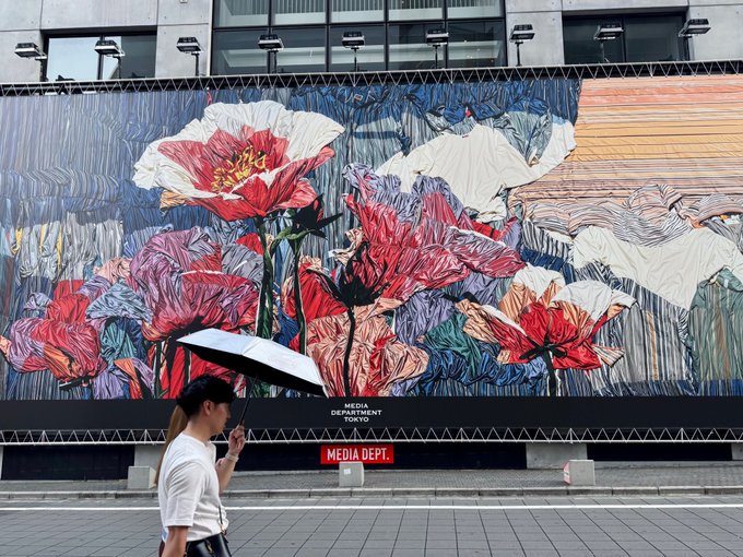 A woman holding a white umbrella walks past a large, colorful floral mural on a building facade labeled "MEDIA DEPT" and "MEDIA DEPT. SHOP." The artwork's oversized blooms and street-level setting create a striking "me and my art" moment, as the passerby becomes a small figure dwarfed by the vibrant botanical scene.