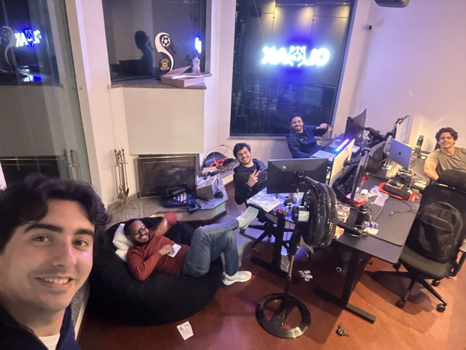 A group selfie of five smiling young men in a tech-focused office/lounge with neon signs, computer monitors, and a bean bag chair.