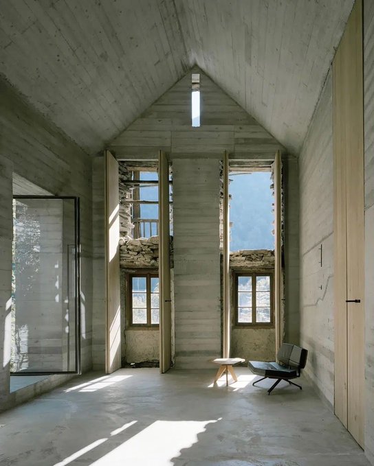 An interior view of a modern concrete house with a tall pitched ceiling, large windows showing a mountain view, a small wooden stool, and a grey chair.