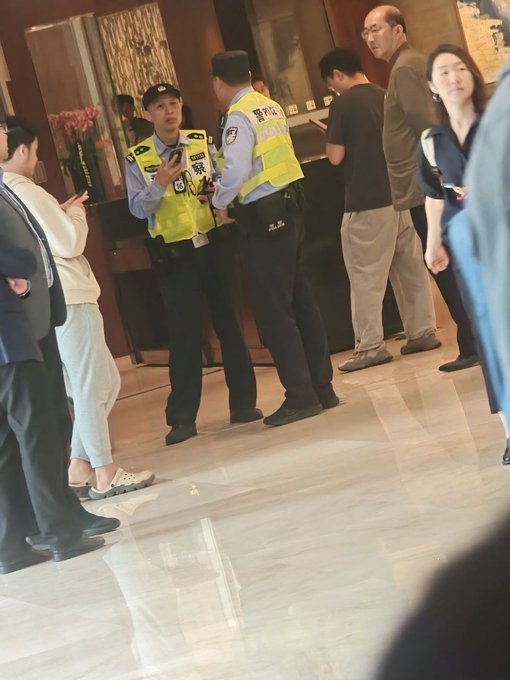 Two police officers in yellow vests stand inside a lobby talking, with several other people in business attire standing nearby.
