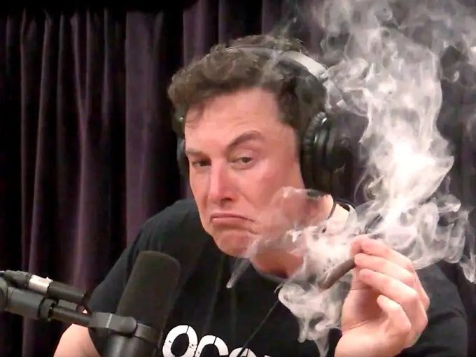 Elon Musk wears headphones and a black t-shirt while smoking a cigar in front of a microphone, with smoke billowing around his face.