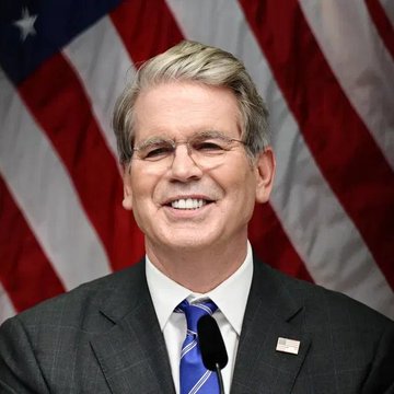 A man with gray hair, wearing a dark suit and a blue striped tie, smiles at the camera with an American flag displayed behind him.