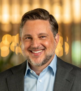 A professional headshot of a smiling middle-aged man with grey hair and beard, wearing a grey suit jacket over a light blue shirt, set against a blurred warm background.