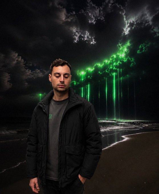 The image features a young man standing alone at the edge of a beach, facing away from the camera while wearing a black jacket that blends with the dark sky above him. His hair is styled in a casual manner, giving off a relaxed vibe. The backdrop consists of a night sky filled with vibrant green streaks coming out of the clouds, creating an intriguing and surreal atmosphere. This image seems to be part of a promotional campaign for a video or digital project related to the artist "@okx". The focus is on the man's presence in the scene, capturing his individuality amidst the stunning visual elements.