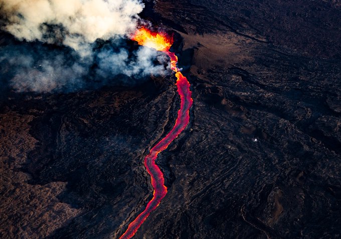An aerial view shows a bright orange lava flow cutting through dark, hardened volcanic rock, with smoke rising from the eruption site.