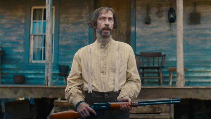 A bearded man with unkempt hair stands on the porch of a weathered blue wooden house, wearing a yellow button-up shirt with suspenders and holding a lever-action rifle, fitting the gritty, rural tone of the post praising the film *Old Henry*.