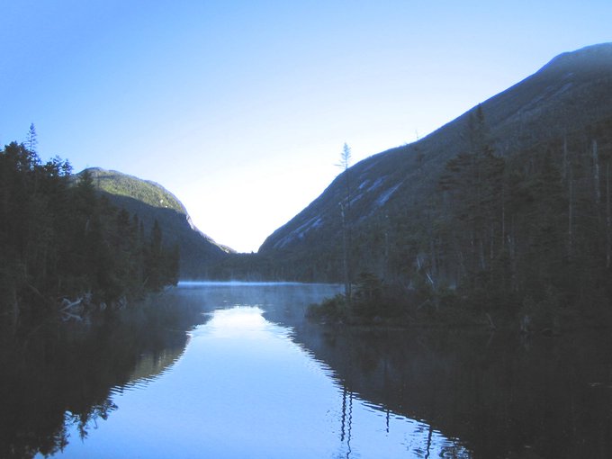 A serene mountain lake surrounded by forested hills under a clear blue sky, with a thin layer of mist hovering above the calm, reflective water surface.