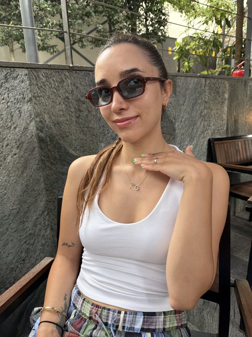 A young woman with brown hair wears sunglasses, a white tank top, and a necklace, sitting outdoors near a concrete wall.