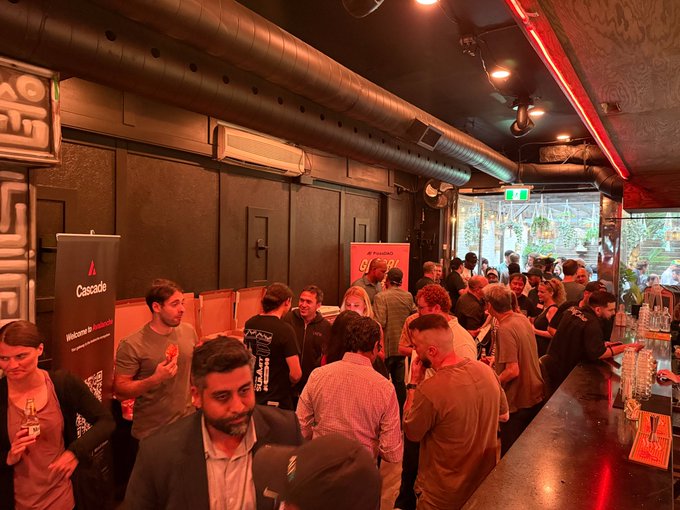 A crowd of attendees mingles in a dimly lit venue with exposed pipes and red neon lighting, featuring visible branding for "Cascade" and "Pizza DAO" on banners and a "Team 1" shirt.
