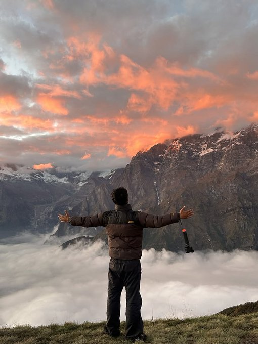 A man stands triumphantly atop a mountain with arms outstretched, surrounded by a sea of clouds and a dramatic pink and orange sunset, appearing carefree despite the caption suggesting otherwise.