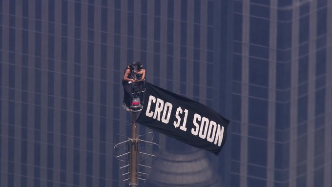 A person rappels down the side of a skyscraper, unfurling a large black banner that reads "CRO $1 SOON," symbolizing $CRO holders' bold declaration of their bullish price target by displaying it prominently atop a city building.