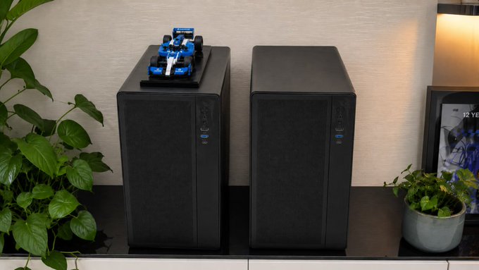 Two black desktop computer towers sit side by side on a dark shelf, with a small blue and white model Formula 1 race car placed on top of the left tower, flanked by green potted plants.